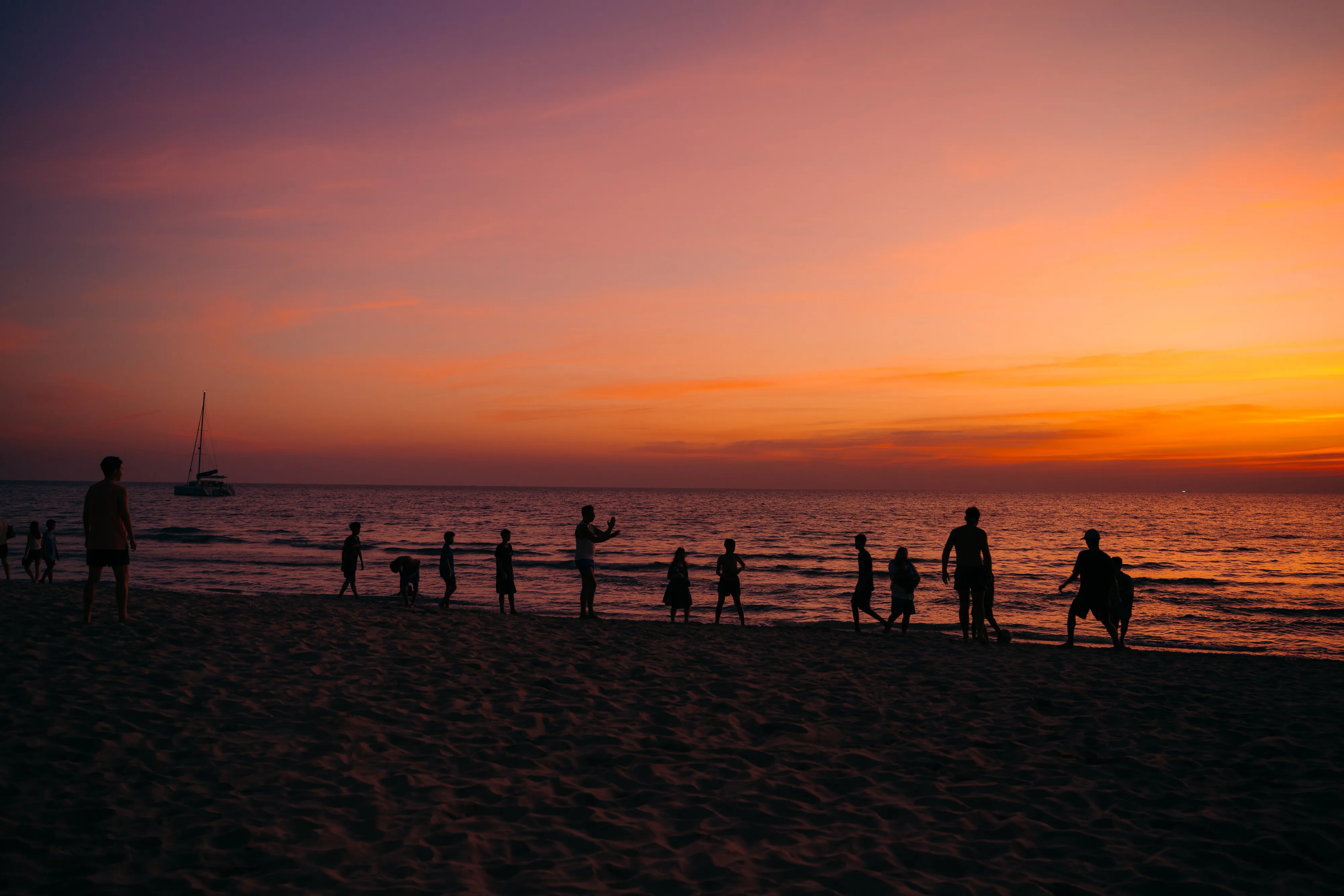 People on a beach at sunset, silhouetted against an orange sky, with a sailing boat on the sea